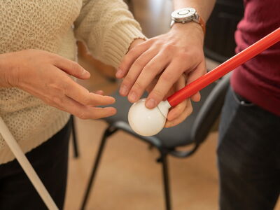 In einer Beratungssituation zu Hilfsmitteln für Blinde und Sehbehinderte ertasten zwei Personen mit ihren Händen die Kugelspitze eines Blindenlangstocks.