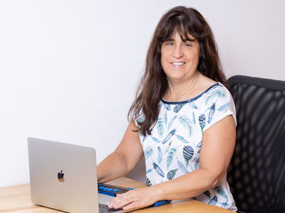 Eine blinde Frau sitzt an einem Schreibtisch und arbeitet an einem Laptop; vor ihr liegt eine Braillezeile, sie blickt lächelnd in Richtung Kamera.