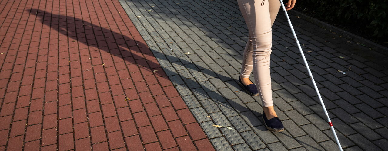 Eine Person geht mit einem Blindenlangstock auf einem gepflasterten Gehweg. Neben ihr ist ein Leitstreifen zu sehen, an dem sie sich orientiert.