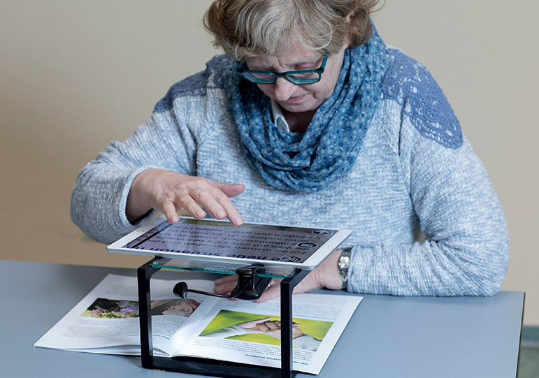 Eine sehbehinderte Frau in hellblauem Pullover mit Halstuch und brille verwendet eine Vergrößerungshilfe, um eine Broschüre zu lesen.