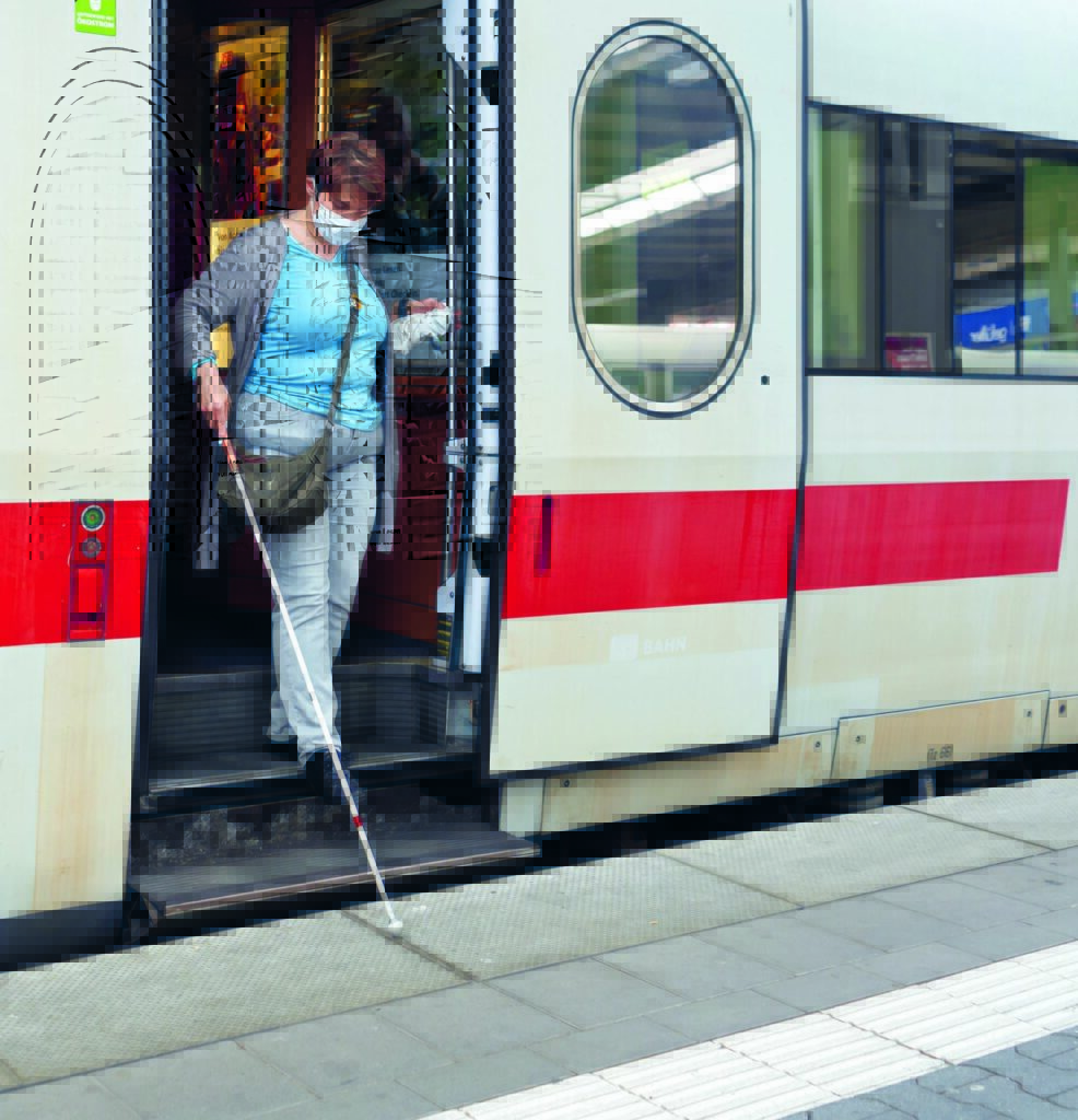 Eine sehbehinderte Frau steigt vorsichtig die Trittstufen eines ICE-Wagens herab. In der rechten Hand hält sie einen Langstock und tastet damit über den Bahnsteig.