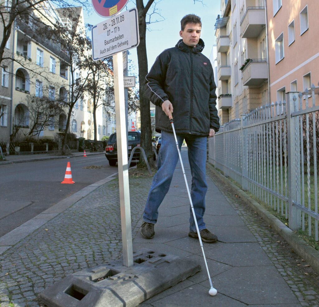 Ein blinder Mann geht mithilfe eines Langstocks einen Bürgersteig entlang. Sein Weg wird durch ein Baustellenwarnschild mit stabilem Sockel unterbrochen, das eine erhebliche Behinderung darstellt.