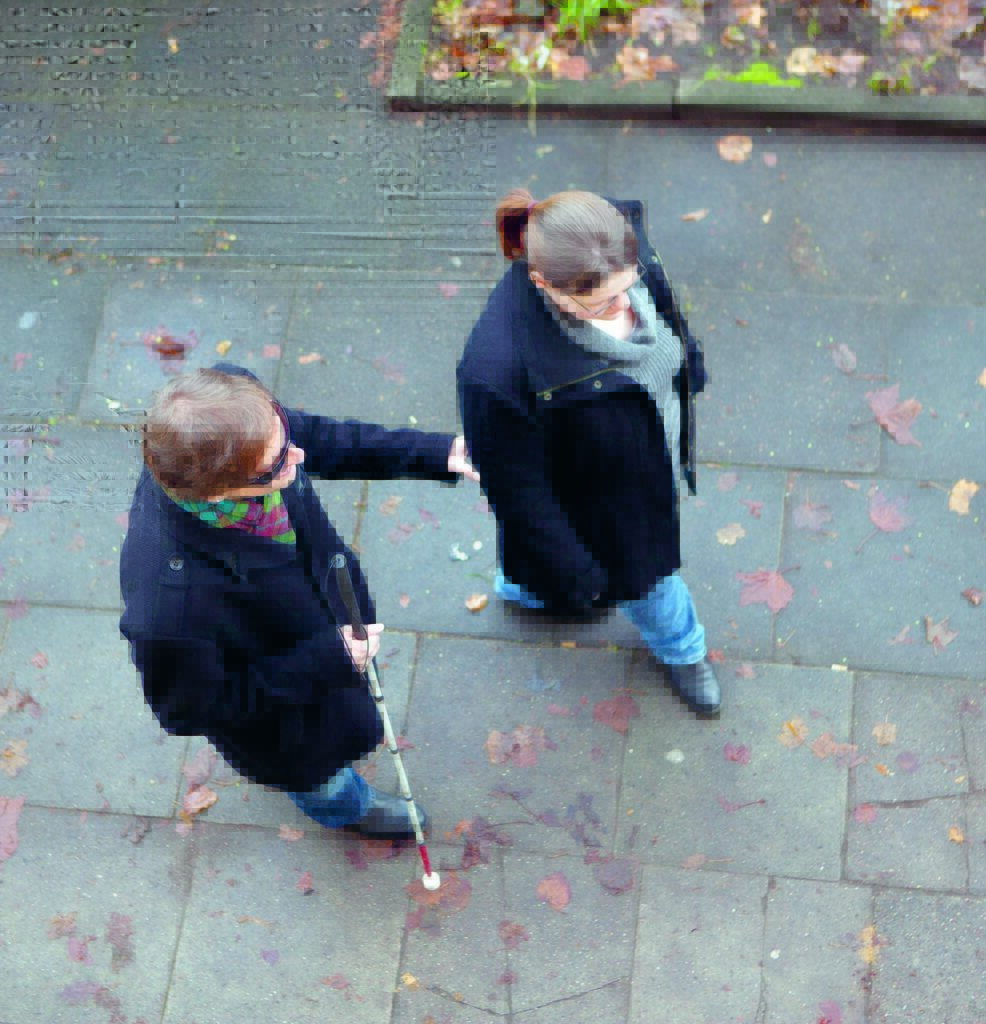 Blick von oben auf zwei Spaziergänger: Eine blinde Person hält sich mit der linken Hand am rechten Arm ihrer sehenden Begleitung fest.