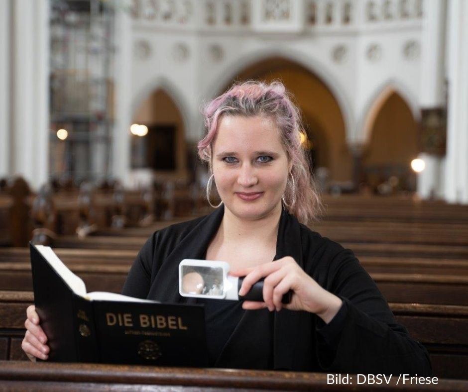 Eine Frau Mitte 20 sitzt in einer Kirchenbank und lächelt in die Kamera, in einer Hand eine Leuchtlupe, in der anderen eine Bibel. Sie hat langes, dunkelblondes Haar mit rötlichen Strähnchen und trägt große Ohrringe. Bildnachweis "DBSV/Friese"