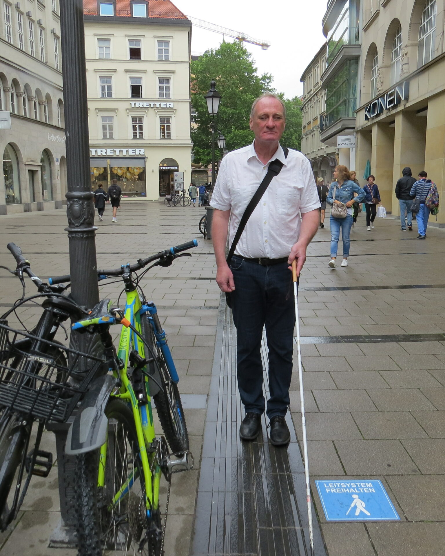 Ein Mann mit Gehstock steht auf dem Leitsystem in der Sendlinderstr. in München. Rechts auf dem Boden ist ein blauer Aufkleber mit Aufschrift "Leitsystem freihalten". Hintergrund: Geschäftshäuser Sendlingerstraße.