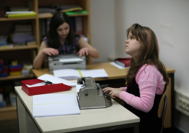 Ein blindes Mädchen in schwarzem Oberteil und rosa Jäckchen schreibt, am Schulschreibtsich sitzend, auf der Braille-Schreibmaschine. Im Hintegrund ein weiteres Kind, das ebenfalls auf der Braille-Schreibmaschine schreibt. Beide haben jeweils auch ein aufgeklapptes Buch in Blindenschrift vor sich liegen.