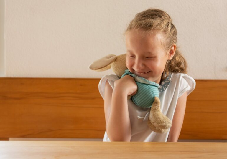 Ein kleines blindes Mädchen mit seinem Kuscheltier, einem Stoffhasen, sitzt an einem Tisch und drückt den Hasen liebevoll an sich.