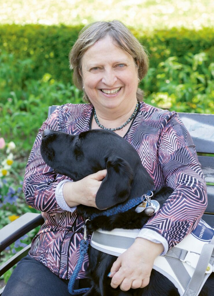 Martina Hellriegel mit führhund Raven auf einer Parkbank sitzend. Raven hat seine Vorderpfoten auf dem Schoß seines Frauchens – dieses legt die Arme um den Hund und lacht.