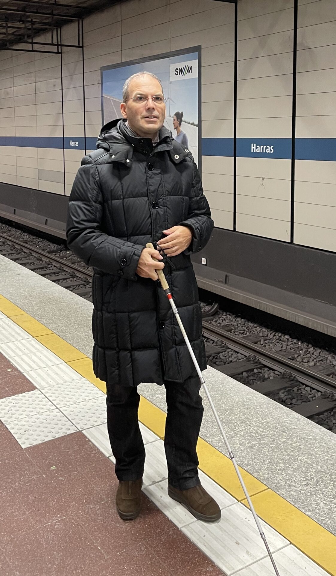 Dr. Stefan Insam steht am Gleis auf dem Leitsysteem am Münchner U-Bahnhof Harras. Er trägt dunkle Winterkleidung und hält in der rechten Hand einen Langstock.