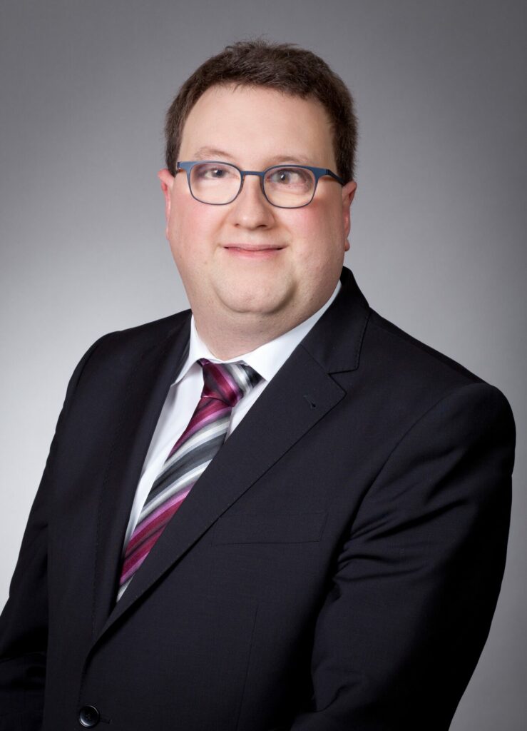 Tobias Michl, Mitglied im erweiterten Vorstand des BBSB e. V., im schwarzen Anzug vor grauem Hintergrund.