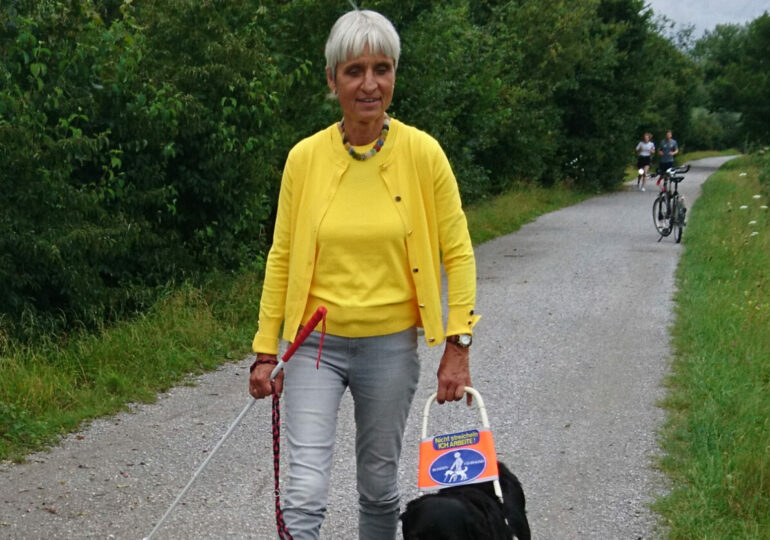 Dr. Eva-Maria Glofke-Schulz in grauen Jeans und gelbem Twinset beim Spaziergang mit ihrem Blindenführhund Max, der sein Geschirr trägt, auf einem Weg inmitten von Grün.